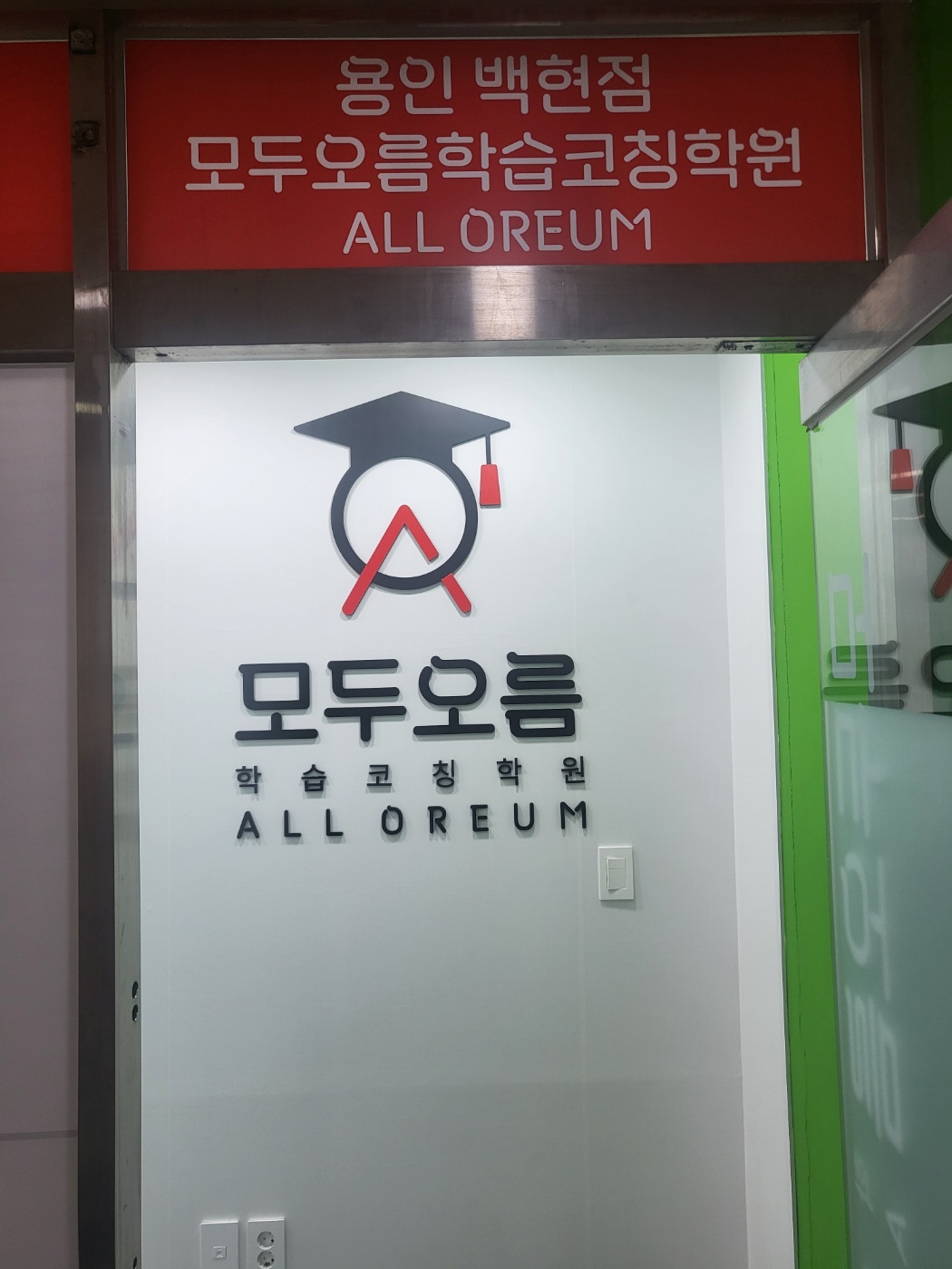용인백현점 입구 — 흰 벽에 학사모를 쓴 원형 로고와 모두오름 학습코칭학원 ALL OREUM 글자가 붙어 있고, 위쪽 빨간 간판에 용인 백현점 모두오름학습코칭학원이라고 적혀 있다