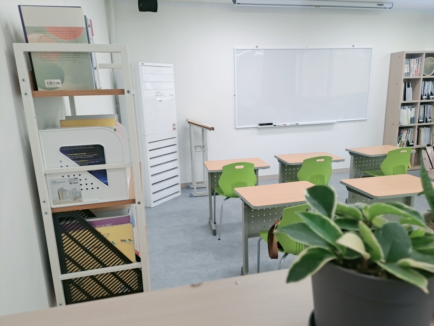 은평점(글로리드) 교실 — 흰 벽에 큰 화이트보드가 걸려 있고 그 앞에 초록색 의자가 딸린 1인용 책상 여섯 개가 놓여 있다. 왼쪽에는 책과 서류함을 꽂은 하얀 선반이, 오른쪽에는 책이 채워진 원목 책장이 있고 앞쪽 책상 위에 초록 화분이 놓여 있다