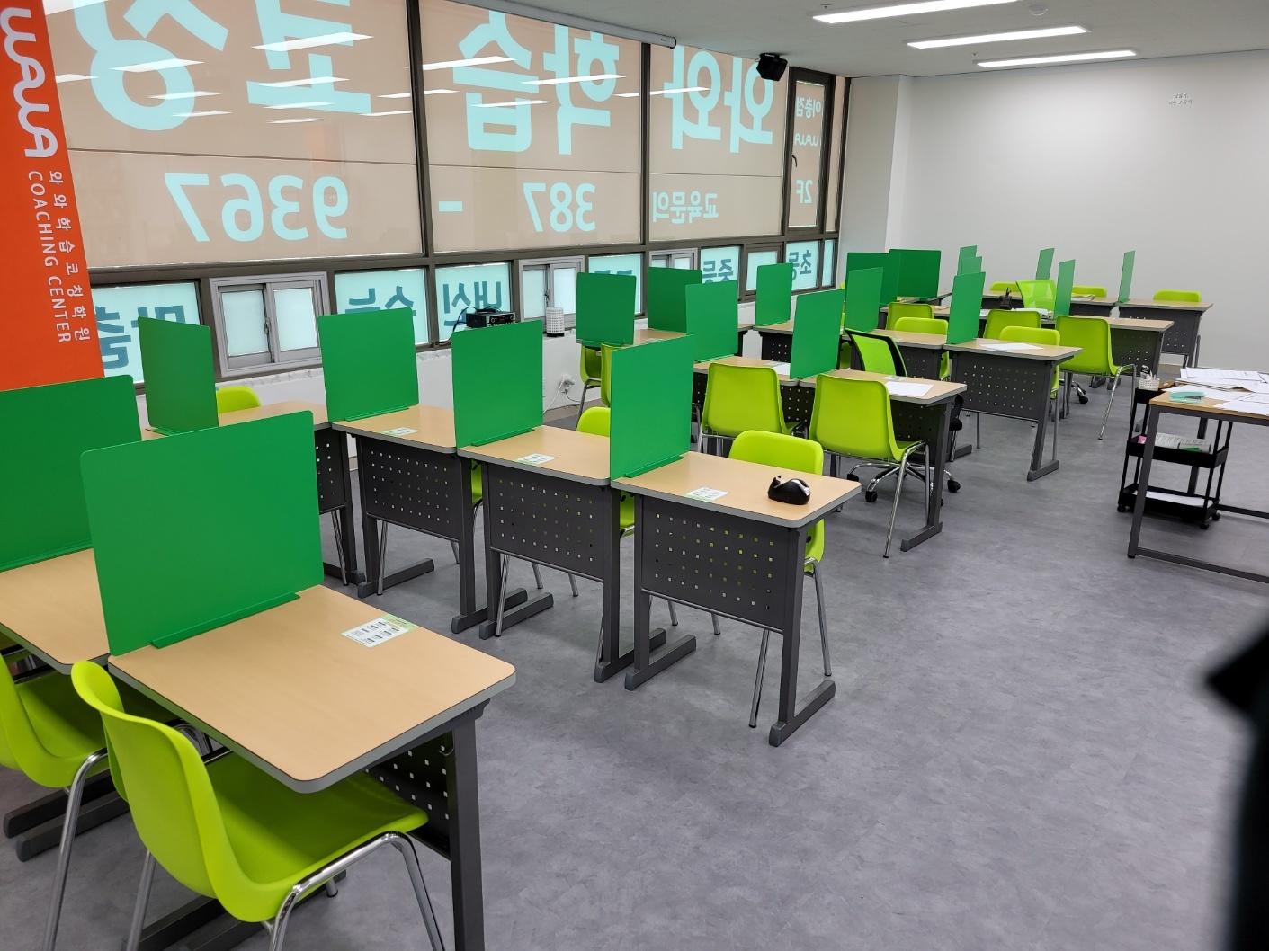 평택 이충동 와와학습코칭 이충점 교실 — 초록 가림판을 세운 책상과 연두색 의자가 여러 줄 놓여 있고, 오른쪽에 코치 책상과 자료를 올린 이동식 카트, 왼쪽 기둥에 주황색 WAWA COACHING CENTER 배너